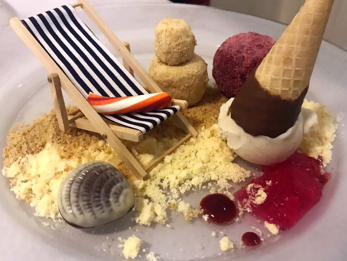 Sweet Flavours by the Sea -Upside down vanilla ice cream with cone on top, shortbread crumb & digestive (sand), stick of rock relaxing on a deck chair, white chocolate rocky road sand castle with mini doughnuts (beach balls) coated in raspberry sugar, chocolate praline shells brushes with white chocolate