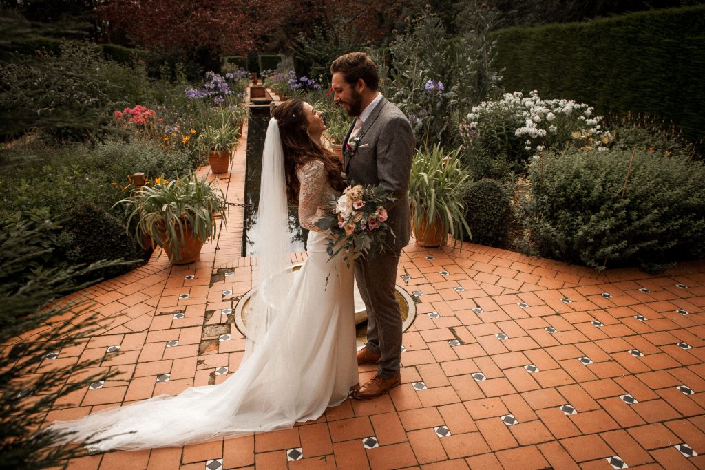 A bride and groom stand facing each other in a garden. The bride holds a bouquet and wears a long white gown with a veil, while the groom is in a gray suit. They are surrounded by lush greenery and colorful flowers on a tiled path.