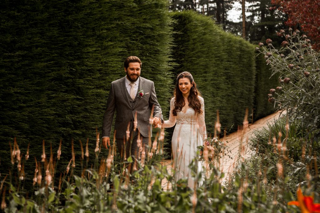 A couple is walking hand in hand down a garden path lined with tall hedges. The man is wearing a gray suit, and the woman is in a white dress. Both are smiling as they walk on the sunny path.