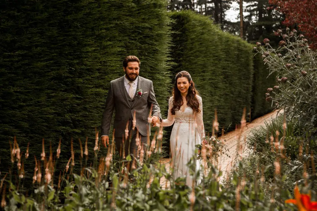A couple is walking hand in hand down a garden path lined with tall hedges. The man is wearing a gray suit, and the woman is in a white dress. Both are smiling as they walk on the sunny path.