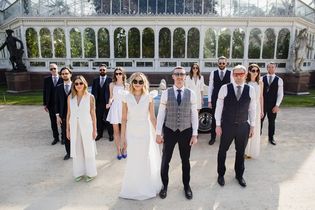 Dine | Exclusive Weddings & Events in Yorkshire | A bride and groom stand confidently with sunglasses, surrounded by their luxury wedding party in stylish suits and dresses. They pose in front of a vintage car and a glass conservatory in a sunny outdoor Yorkshire setting.