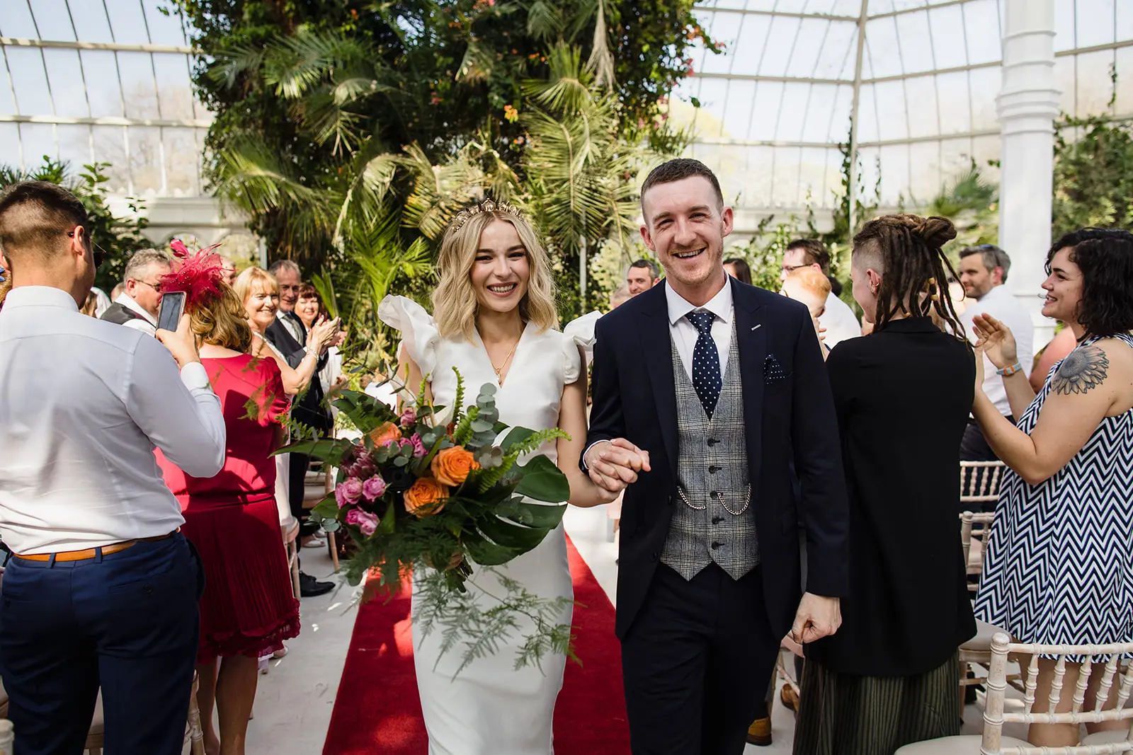 Dine | Exclusive Weddings & Events in Yorkshire | A bride in a white dress and a groom in a suit walk down the aisle at their enchanting Yorkshire wedding, smiling and holding hands. Surrounded by applauding guests amidst lush green plants in the greenhouse, the bride holds a bouquet of flowers, setting the stage for unforgettable events.