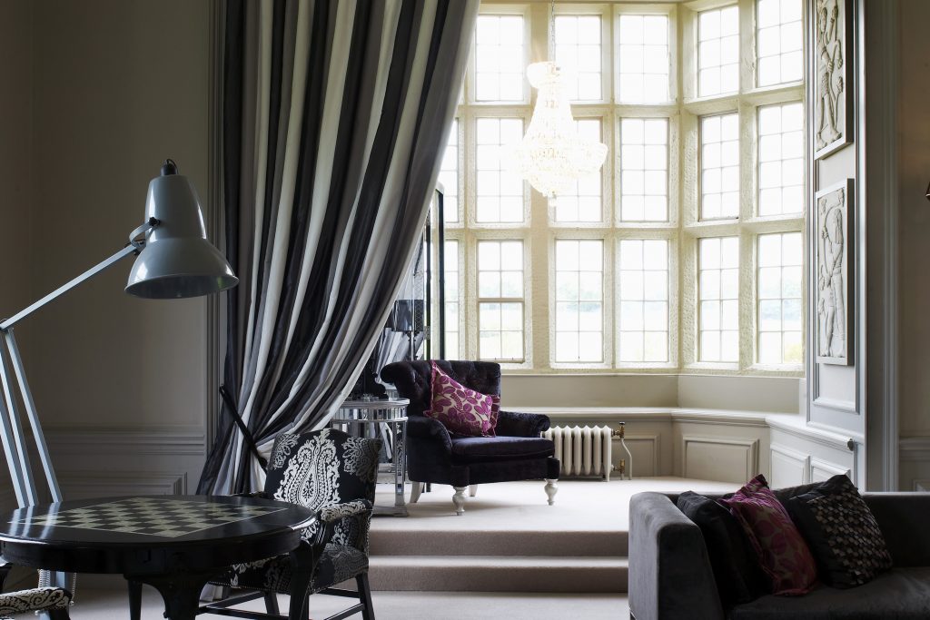 Dine | Exclusive Weddings & Events in Yorkshire | The room exudes luxury and elegance, perfect for hosting intimate events or even a Yorkshire wedding. It boasts a large window, ornate chandelier, and a black-and-white floor lamp. A dark chair with a patterned purple pillow complements the detailed curtains and plush sofa adorned with colorful pillows.