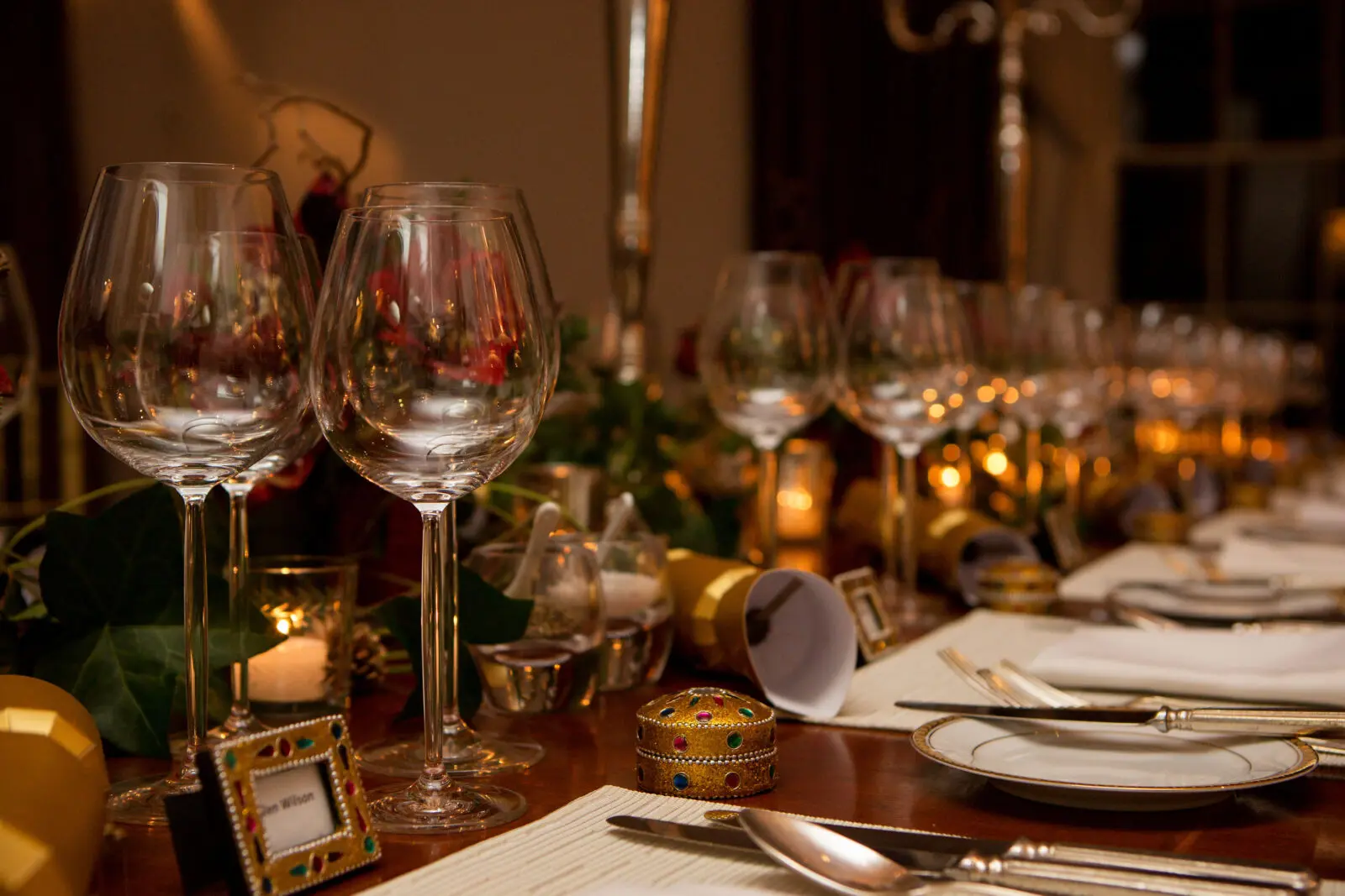 Dine | Exclusive Weddings & Events in Yorkshire | A lavishly set dining table at a grand Yorkshire wedding features several rows of empty wine glasses, ornate name cards, and decorative gold accents. Candles and soft lighting add a warm, festive ambiance. Plates and napkins for the delectable food are neatly arranged by each setting.
