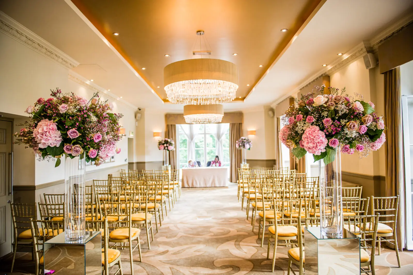 Dine | Exclusive Weddings & Events in Yorkshire | An elegant wedding ceremony room with rows of gold chairs facing a table at the front exudes luxury. The aisle is adorned with tall floral arrangements of pink and purple flowers, while a large chandelier hangs from the ceiling, casting a glow over this exquisite event setting as sunlight streams through the window.